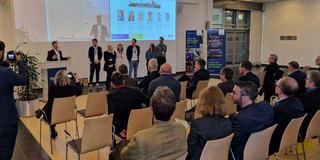 Auf dem Foto ist eine Konferenzsituation zu sehen. Mehrere Menschen befinden sich im vorderen Bereich, wo auch eine Beamerprojektion an der Wand zu sehen ist. Ein Redner stellt 5 Personen vor. Diesen werden von ca 20 sitzenden Personen zugehört.